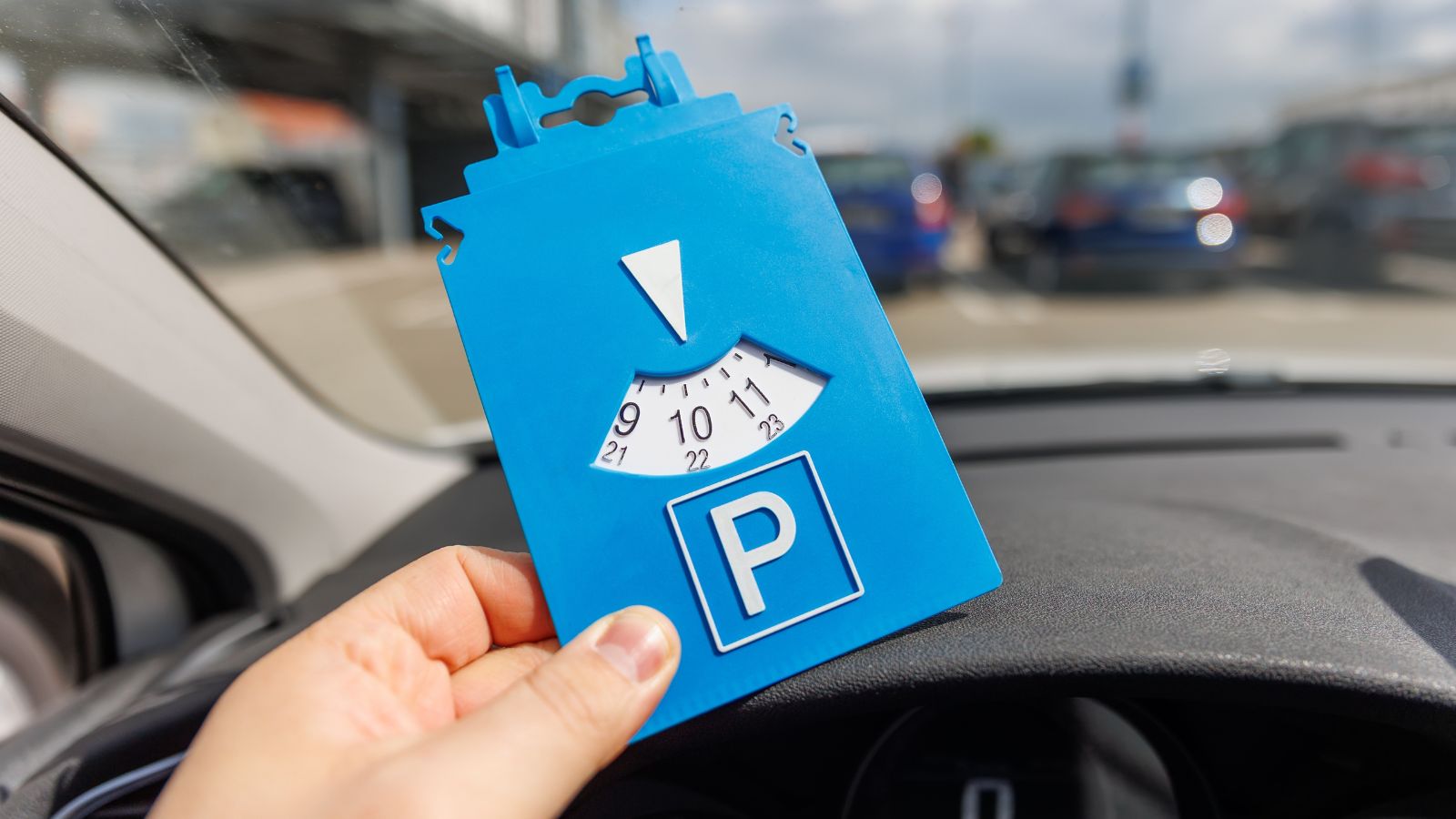 Driver hand hold blue parking disc show arrival time near large shopping complex mall. Parking time restrictions in busy parking lots. Limited time duration park place zone