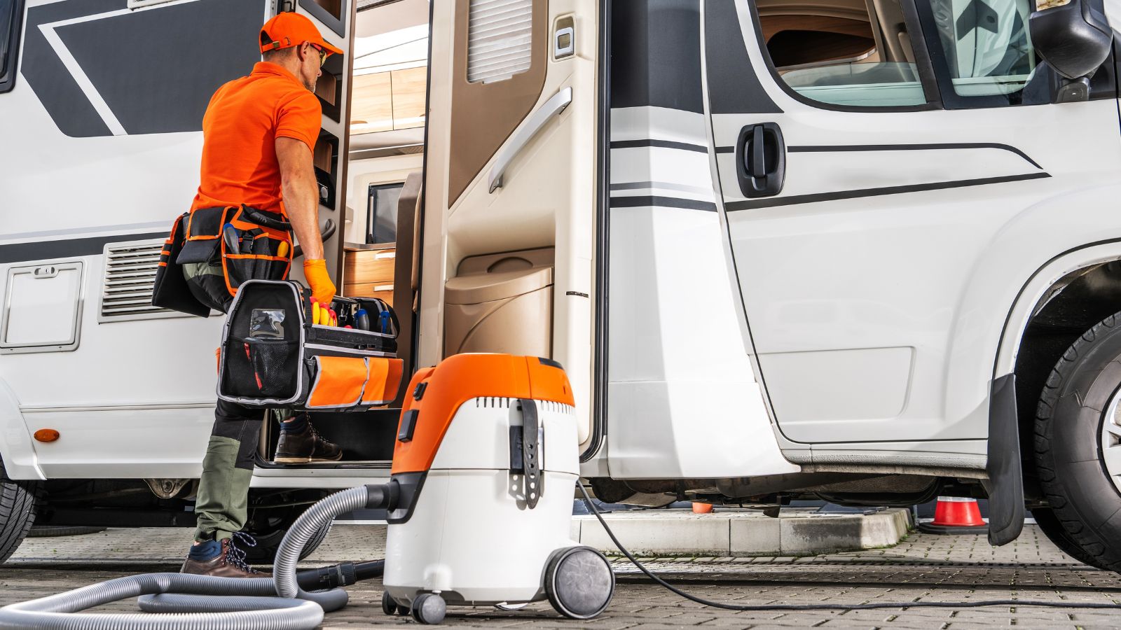 A worker in orange attire uses a vacuum cleaner to clean the interior of a camper van parked outdoors during daylight hours, ensuring thorough maintenance