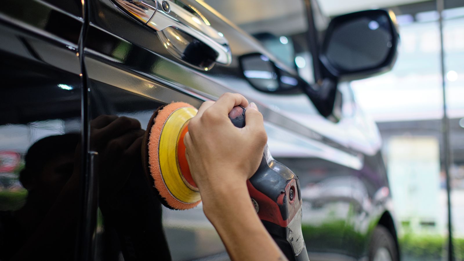 Professional auto detailer hand holding rotary polisher while polishing paint surface of shiny black sedan. Car detailing, car wash, and paint correction concept. Random orbital polisher background.